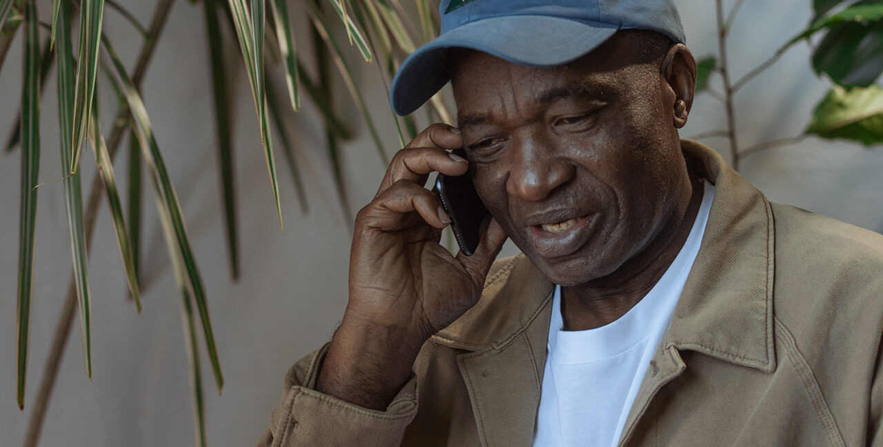 Older man in brown jacket wearing a blue cap talking on a mobile phone.