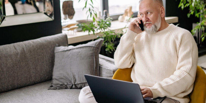 Image shows a man with a beard sat on a sofa with a laptop on his knee. He is wearing a light coloured jumper and holding a mobile phone up to his ear staring into the distance.